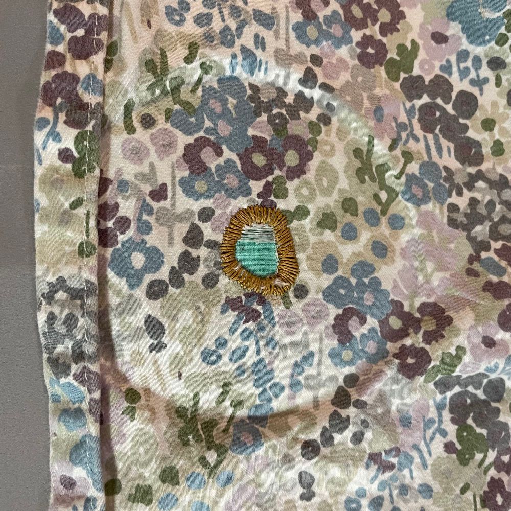 The front of the sheet with the hole mended, and the embroidery hoop removed, you can see the gold thread has been satin stitched around the open hole, revealing a green center.