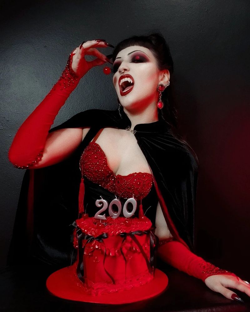 Sou eu, toda vampirona, comemorando meu aniversário! Tenho franja curtinha, maquiagem pesada e sobrancelhas finas, além de unhas muito longas. Estou toda vestida e maquiada de preto e vermelho com muito strass e pedraria, com um vestido preto de veludo, capa preta de forro vermelho e sutiã à mostra em vermelho com tassels e pedraria, luvas também vermelhas com pedraria. Estou combinando com meu bolo que também é preto e vermelho e tem velinhas indicando meus 200 anos em cima. Estou deliciosamente comendo uma suposta cereja & minhas presas estão à mostra