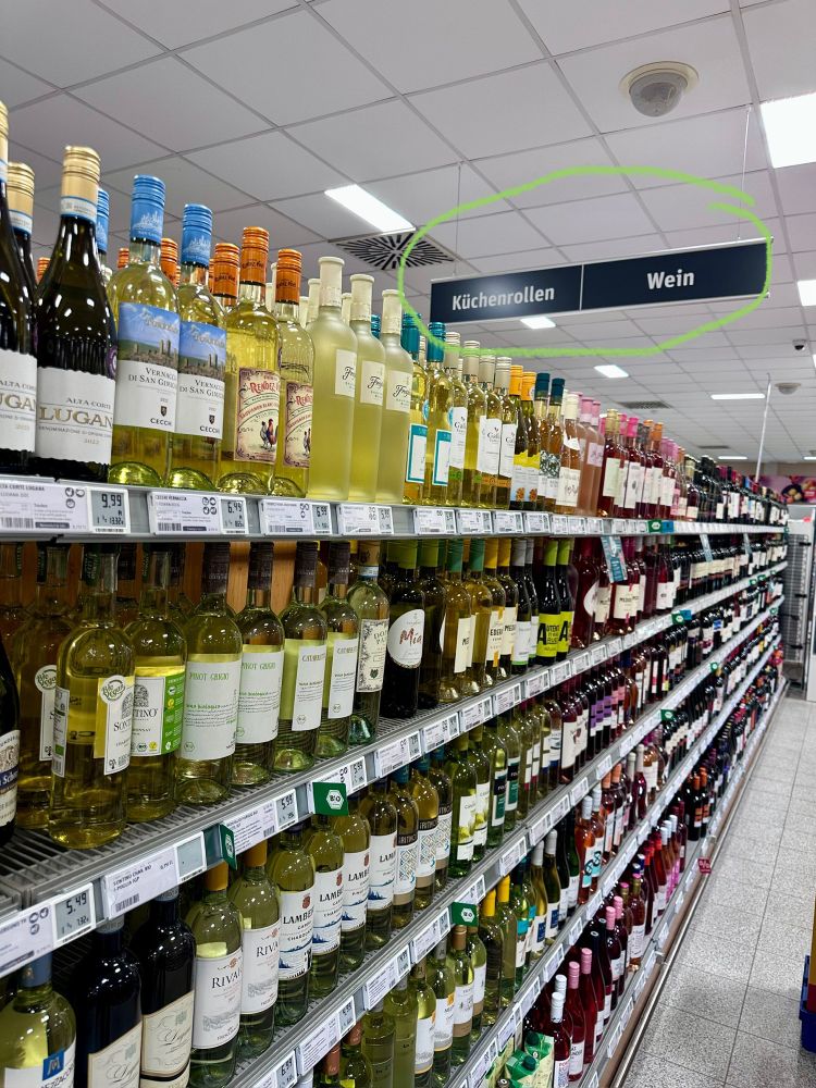 Deutsches Supermarktregal mit hunderten von Weinen (mehrheitlich in Weißglas gefüllt). An der Decke zwei Orientierungsanzeiger: „Wein“, links daneben: „Küchenrollen“. 