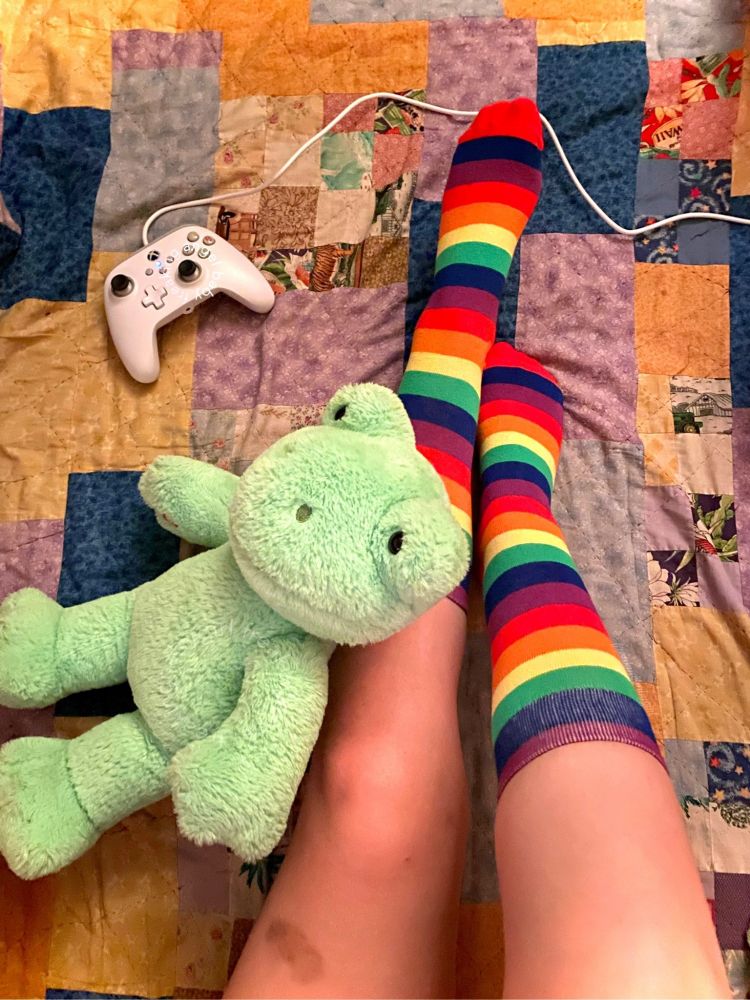 picture of my legs with knee high rainbow socks on, taken from above on a quilted blanket. my right leg is lifted and resting on my left leg is a frog build a bear. further up on the blanket is a white xbox controller. there is a bruise on my thigh.