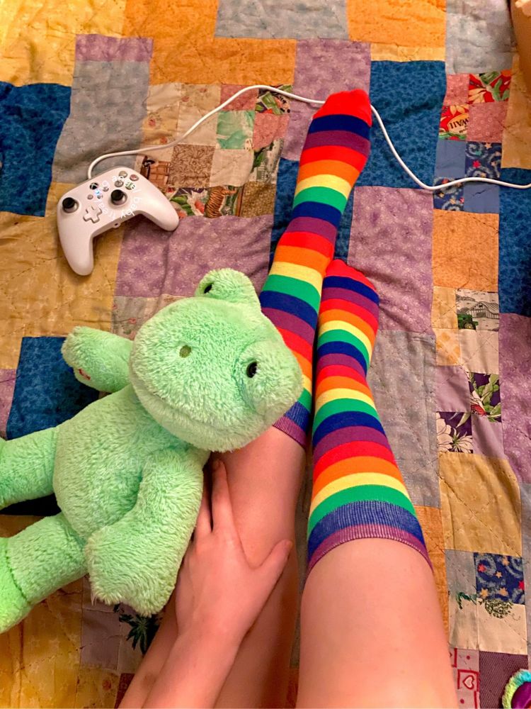 picture of my legs with knee high rainbow socks on, taken from above on a quilted blanket. my right leg is lifted and resting on my left knee is my hand and a frog build a bear. further up on the blanket is a white xbox controller. 
