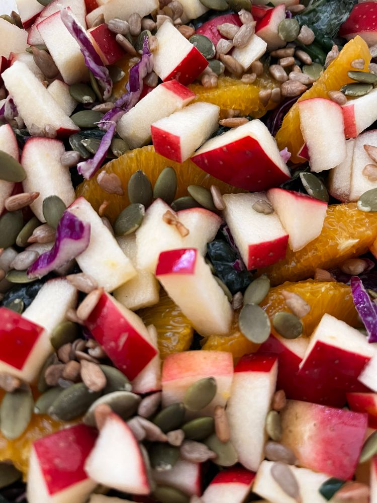 A close up of a salad with orange sections and chopped red apples sitting on a base of kale and purple cabbage. Sunflower and pumpkin seeds are scattered on top.