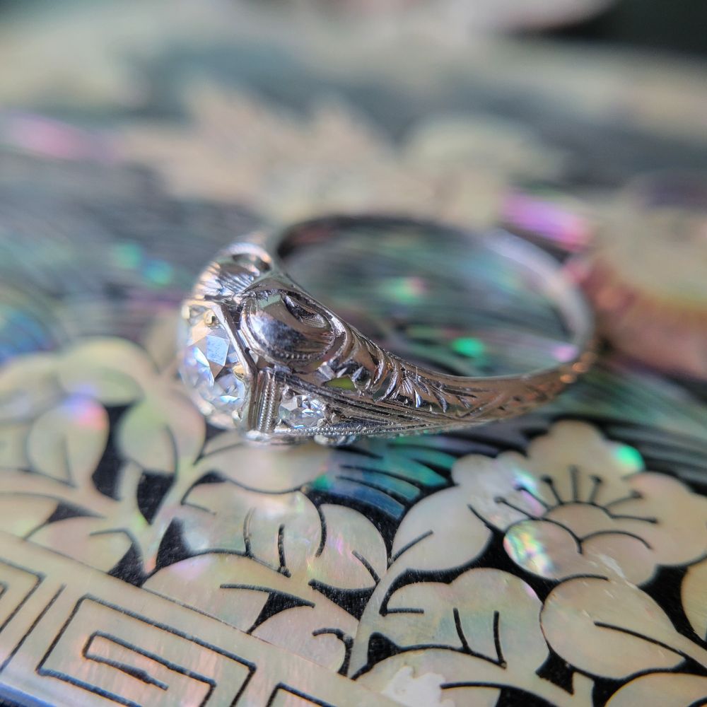 A vintage Art Deco ring in platinum and diamonds is seen in 3/4 view on top of a box inlaid with mother of pearl. The ring features a hexagonal center setting surrounding an old European cut diamond, milligrain, and saw-pierced and engraved Deco motifs, all bathed in ethereal morning sun. 