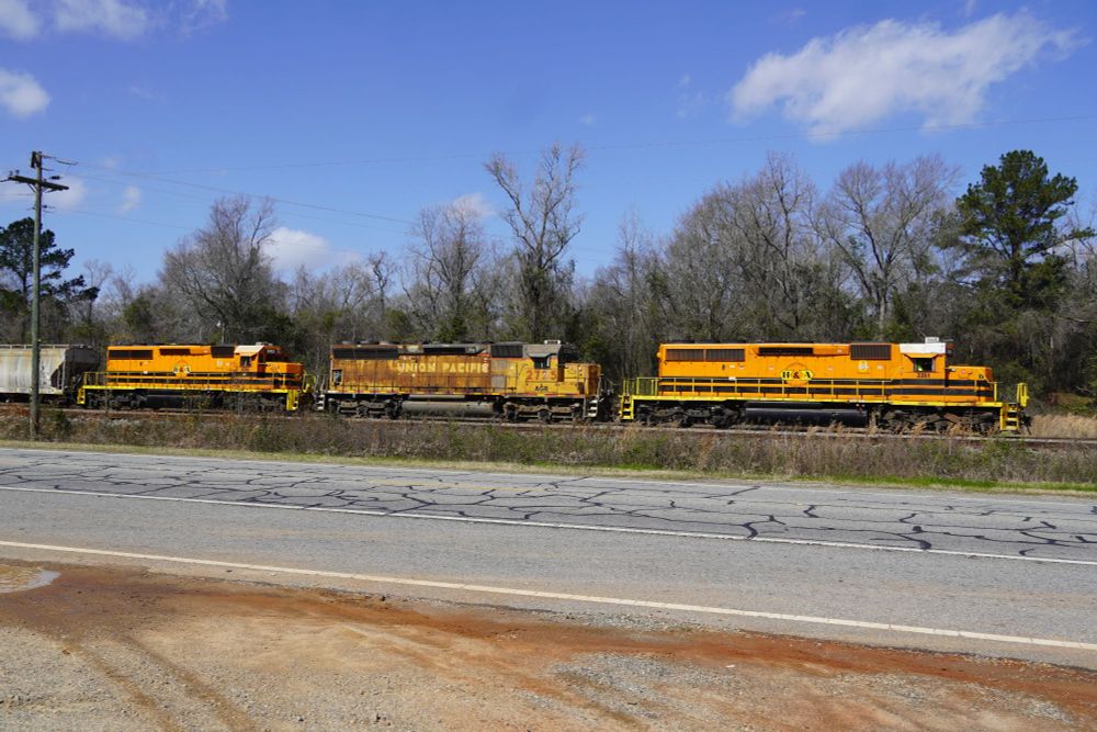 Three diesel locomotives. The sky is blue with a few clouds. Two of the locomotives are Electro-Motive (General Motors) SD-40-2 painted in orange with two black horizontal stripes - the colors of the holding company. The center locomotive is an Electro-Motive SD-45 in gray and yellow of the Union Pacific. The paint is faded and has rust showing through much of the paint.

