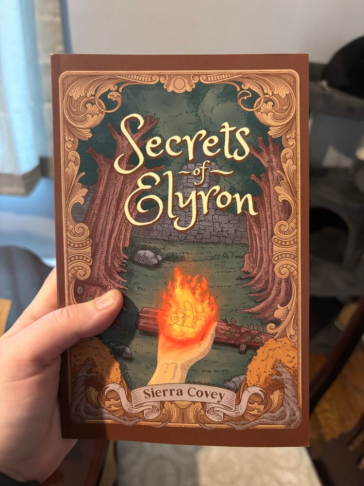 A white hand holding a paperback copy of the book, “Secrets of Elyron,” by Sierra Covey. The cover features the title in gold lettering, a forest in the background, and a white hand holding a flame in the foreground. 