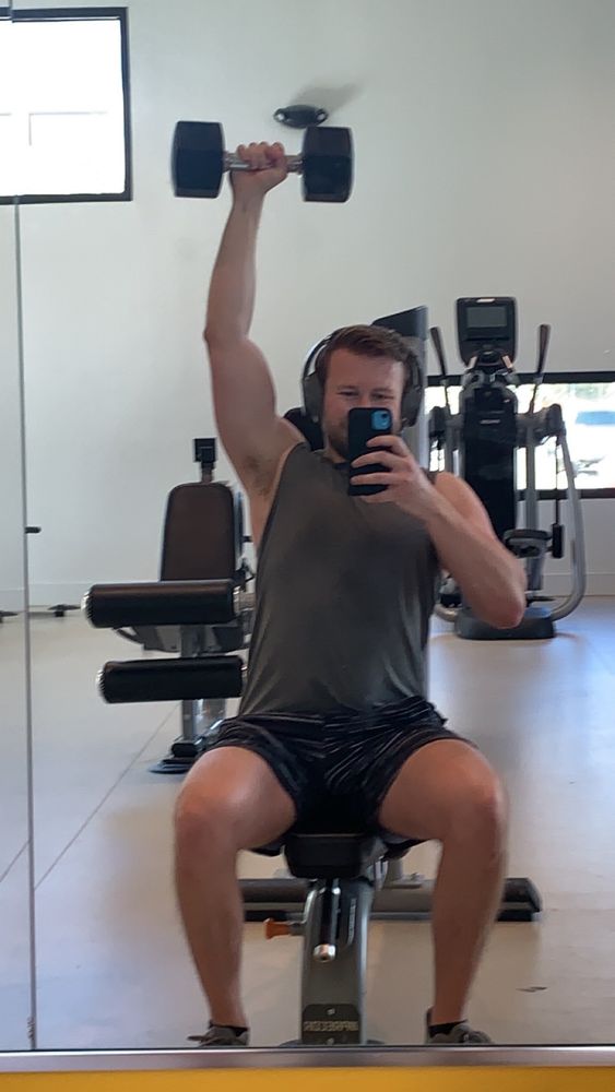 A gym selfie of a man with a dumbbell held over his head showing his pit