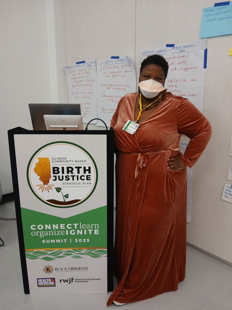 Winifred Burton a fat Black middle aged woman with short silver and black hair, wearing a peach velvet dress and a green and brown necklace stands next to a lecturn with a sign for the 2025 Illinois Community Based Birth Justice Strategic Plan Summit, with some notes from consensus breakout sessions taped up on the wall behind her.