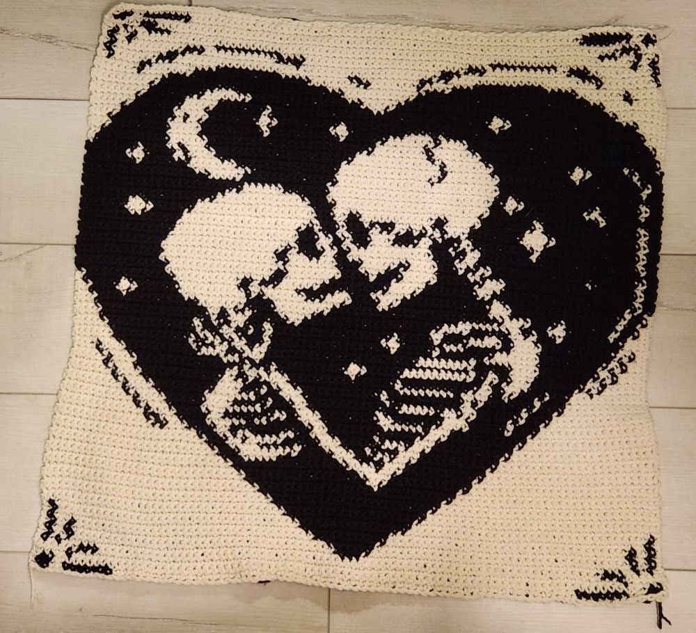 A crocheted blanket sitting on the floor, there's a pattern that looks like two skeletons about to kiss Under the Stars inside of a heart.