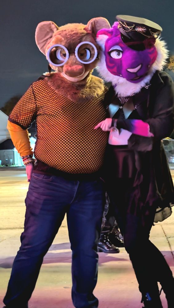 On the left is a man with a brown rat fursuit head on, with large goggles over the eyes. He is wearing a black mesh tshirt over a long sleeve yellow shirt, and jeans. On the right is a woman with a purple tiger fursuit head on and an ornate dominatrix hat over one ear and eye. She is dressed in all black save for a purple/pink arm warmer.