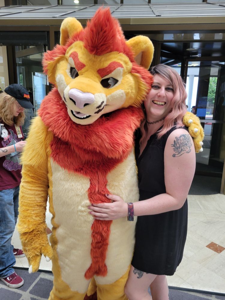 Lion fursuiter and pink-haired girl in a black dress hug for a picture