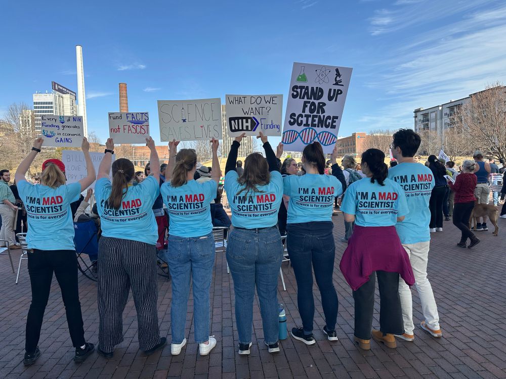 Seven people stand in a row holding up various signs advocating for science. Each person is wearing a blue shirt that reads “I’m a MAD scientist” with a red line under it. Below the red line it reads “Science is for everyone”.