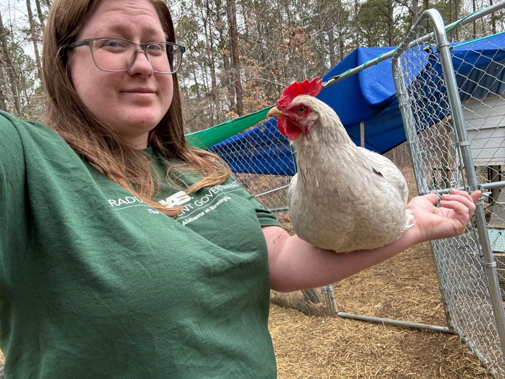 Caitlyn is wearing a green UAB shirt and black glasses. Their hair is down with some tucked behind their ear. On their left arm, a white hen is sitting looking at Caitlyn.