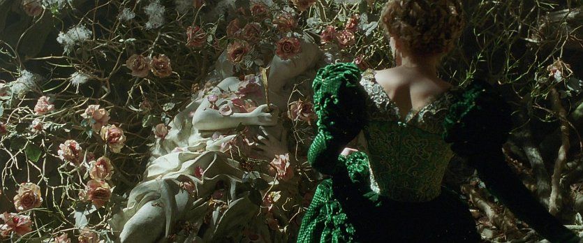 Lea Seydoux in La Belle et la Bete, wearing a green velvet dress and her hair up, seen from the back as she looks at marble status of a dead woman holding an arrow to her chest surrounded by roses.
