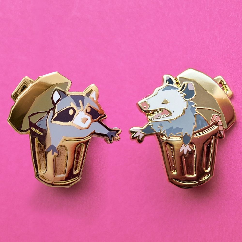 Gold plated enamel pins of a raccoon in a trash can and a possum in a trash can