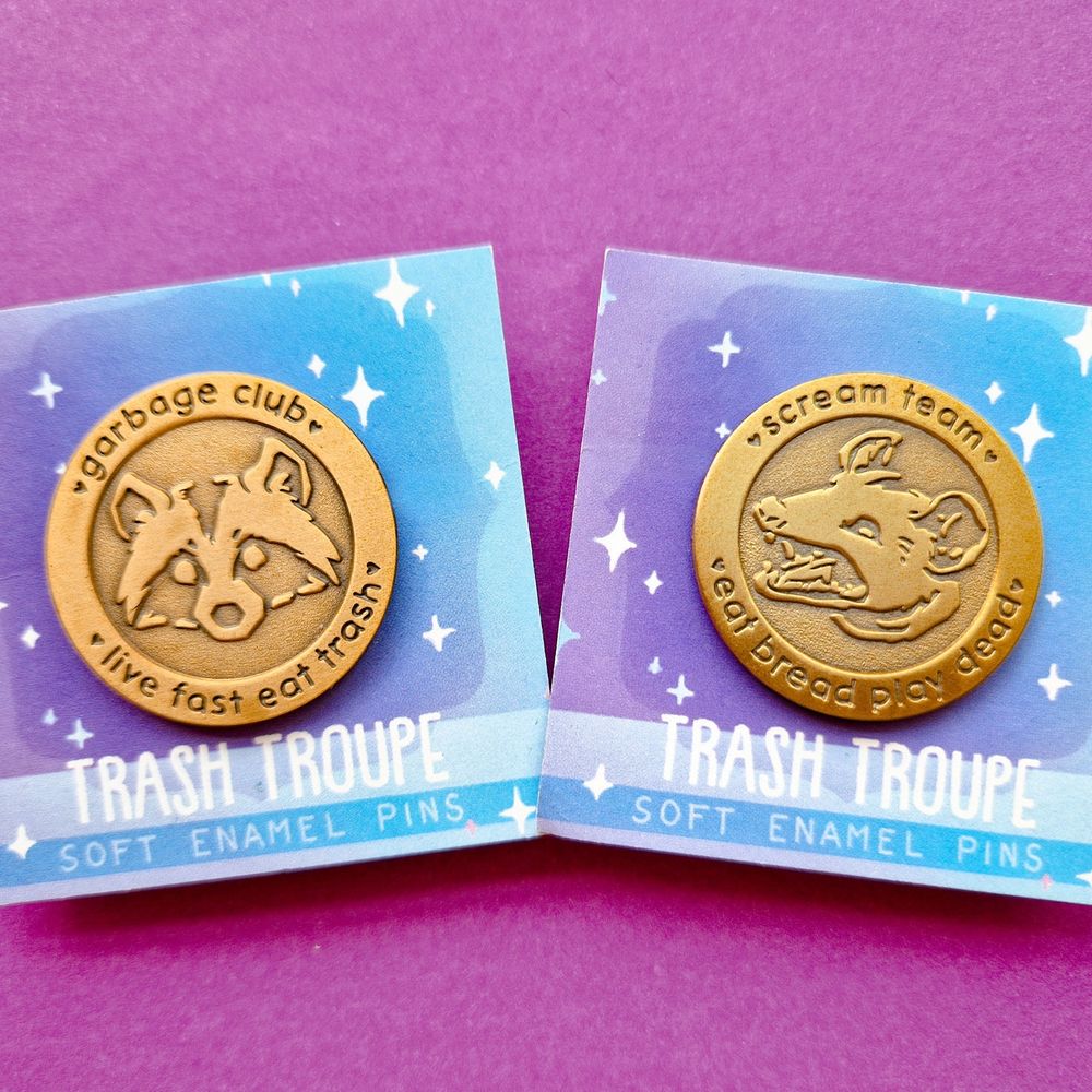 Two antique gold plated enamel pins, the left is a raccoon head with the text 'garbage club, live fast eat trash', the right one is an opossum with the text 'scream team, eat bread play dead' 