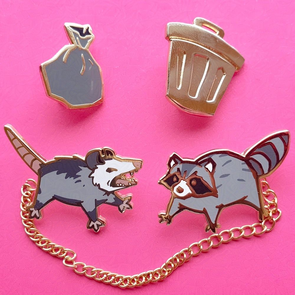 Two collar enamel pins with chain, the trash bag and trash can attachments are pictured above, on the left is a screaming opossum pin and on the right is an angry raccoon pin, they are attached by a golden chain
