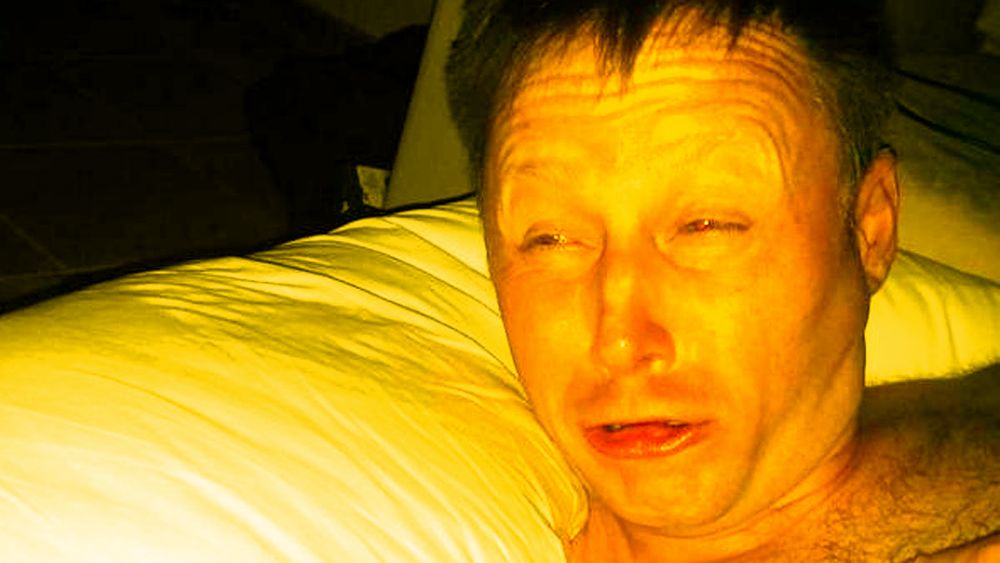 A man in bed squinting as he is bathed in yellow light.