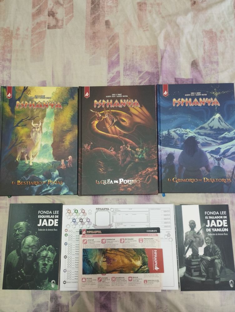Los tres libros del juego de rol Isphanya (el bestiario, la guía y el grimorio), junto con hojas de personaje y tarjetones. Flanqueando, los libros «Esquirlas de jade» y «El tallador de jade de Yanlun», de Fonda Lee.