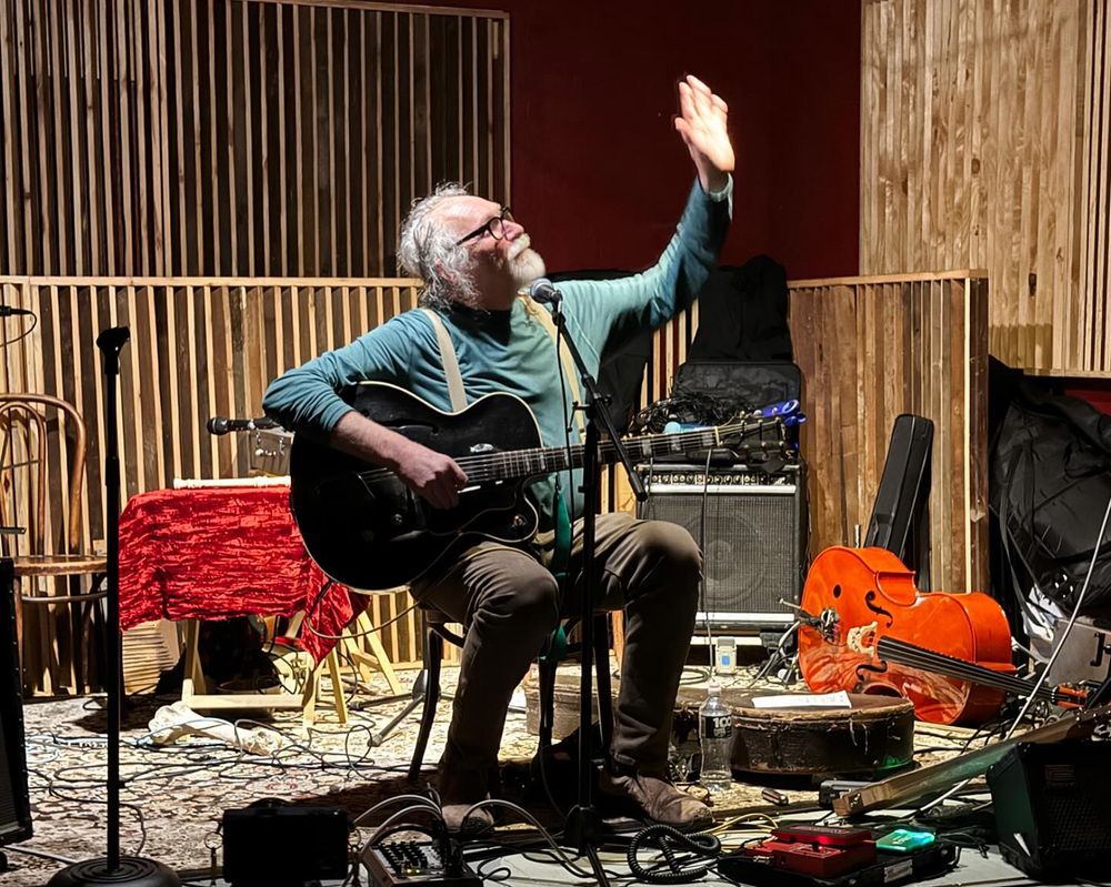 Bill Brovold seated on stage with a guitar and many other instruments around him on the floor