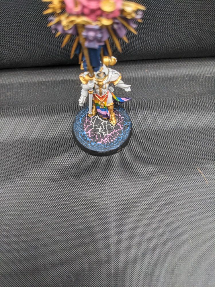 Eagle Eyed view of a Sisters of Battle Imagifier painted in White and Gold, with Pride coloured robes and a Bisexual coloured Icon. The base is made with crackle paint, with the colours of the Trans flag beneath the cracks 