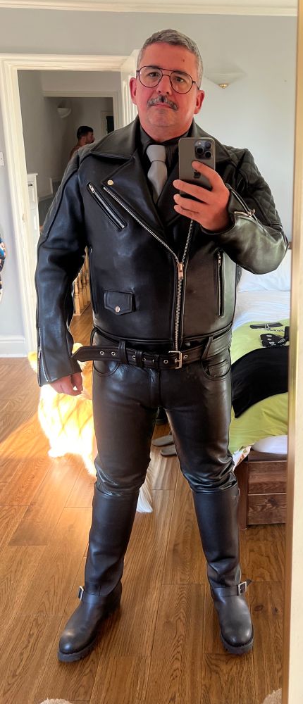 Picture of a man in full leather (tall boots, jeans, shirt, grey tie) with a classic biker leather jacket. 