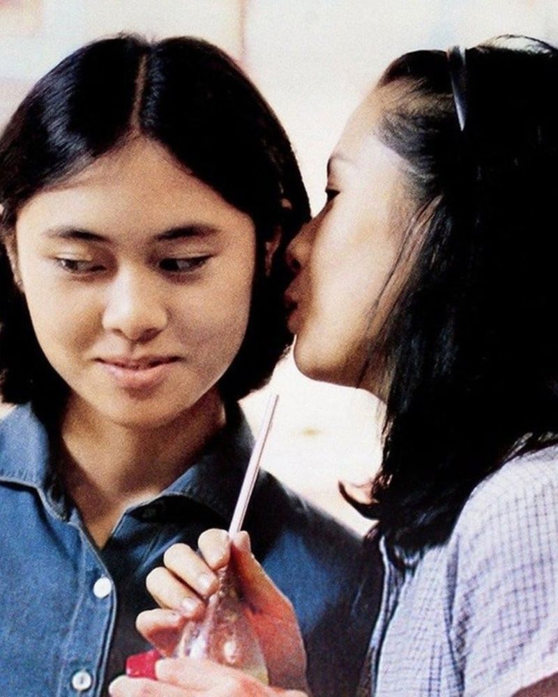 Two young adults share a moment, one holding a drink with a straw while leaning closer to the other, set against a bright backdrop.


