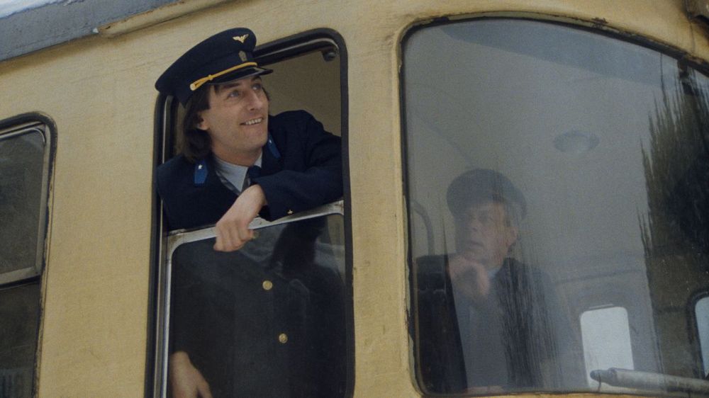 A young train driver looks out of pale yellow train locomotive smiling whilst his colleague looks at him