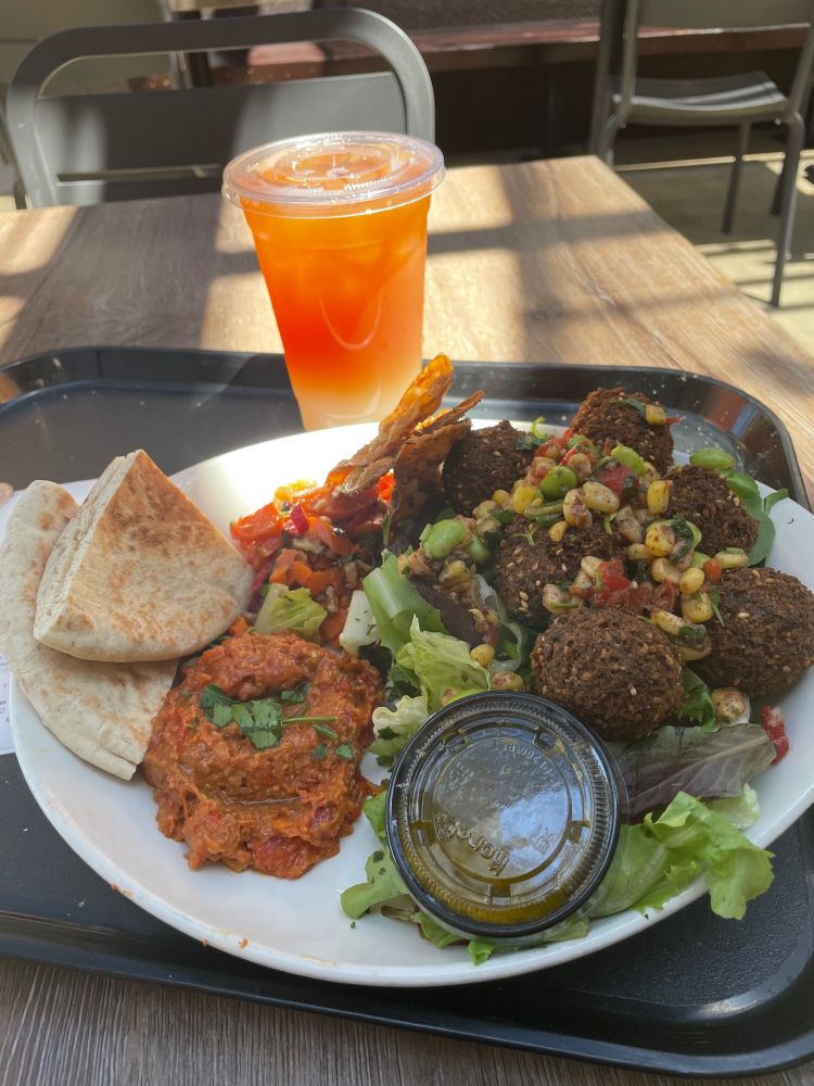 A plate with pita bread, roasted red pepper dip, carrots with walnuts, and a falafel salad. A cup of Arnold Palmer (iced tea and lemonade) on the tray behind the plate.