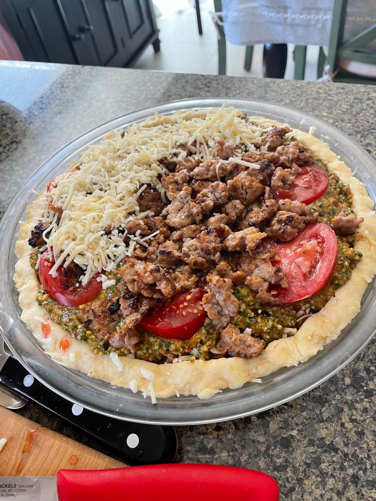 Tomato pie during assembly. You can see layers of pesto, tomato, sausage, and grated cheese. 