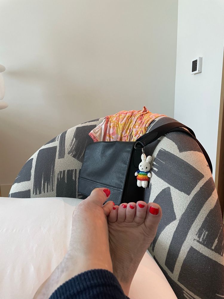 Closeup of my feet with fire-engine red toe nails on a neatly made hotel bed. My favorite black Coach purse with its crochet bunny in the background. 