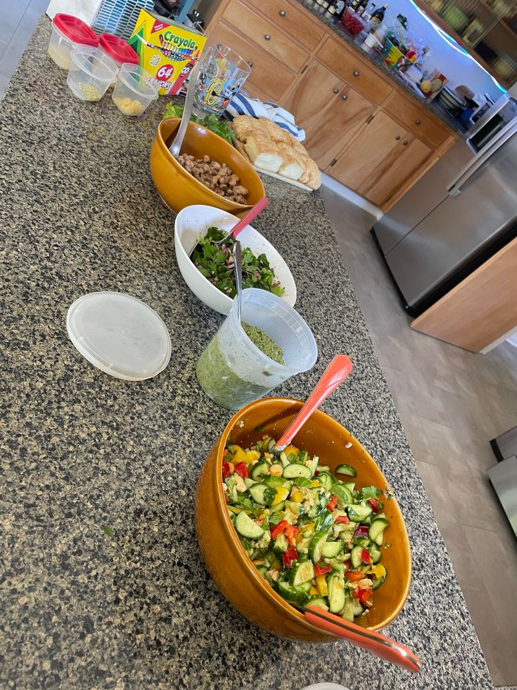 Lined up on a kitchen counter: a bowl of cucumber salad, a container of pesto, an herby salad, and beans. 