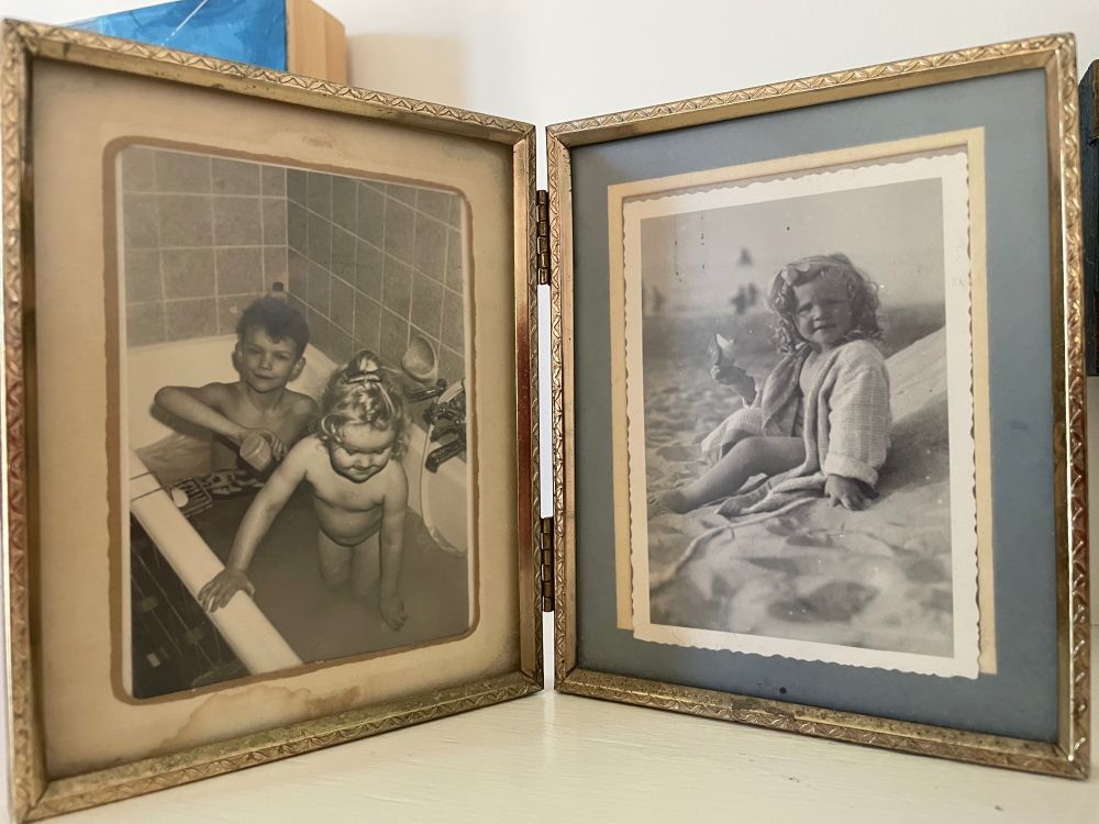 a hinged frame with two black and white photos: on the left, a boy child and a girl child in a bathtub. On the right, a girl child sitting on a beach, her shoe in one hand. 