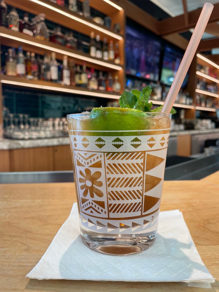 Closeup of a tiki glass with a rum drink on a bar counter. Shelves full of bottles in the background. 