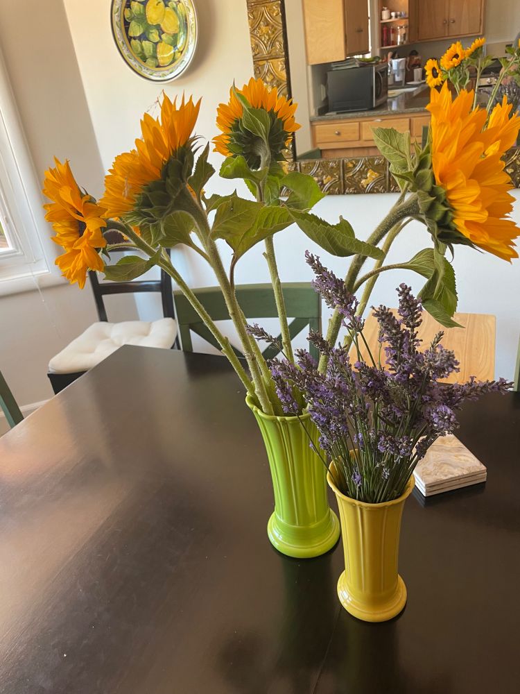 A larger green vase with a half dozen sunflowers, and a smaller yellow vase with a bunch of lavender, on a black dining table. 