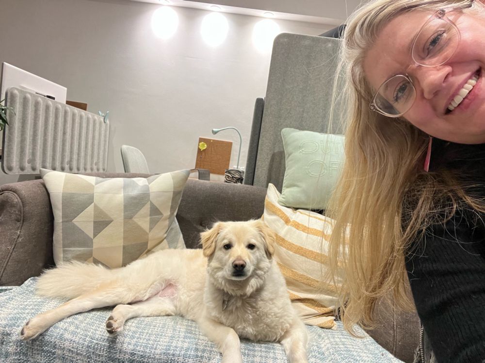 A selfie of Jen smiling on the right with cookie, a small white rescue dog’ sat on the sofa looking at the camera