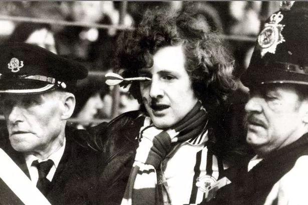 Football fan with a dart sticking out of the bridge of his nose with police taking him from the ground