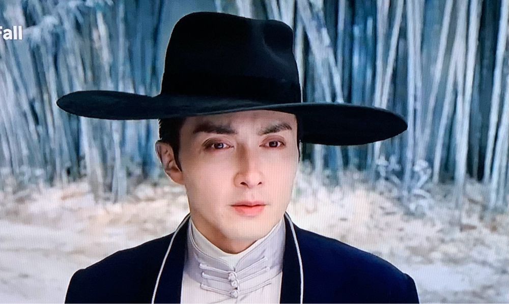 Our broad shouldered pale faced vampire dress in black and white in front on snow covered bamboo. Wearing an exaggeratedly broad brimmed floppy hat generally worn by women