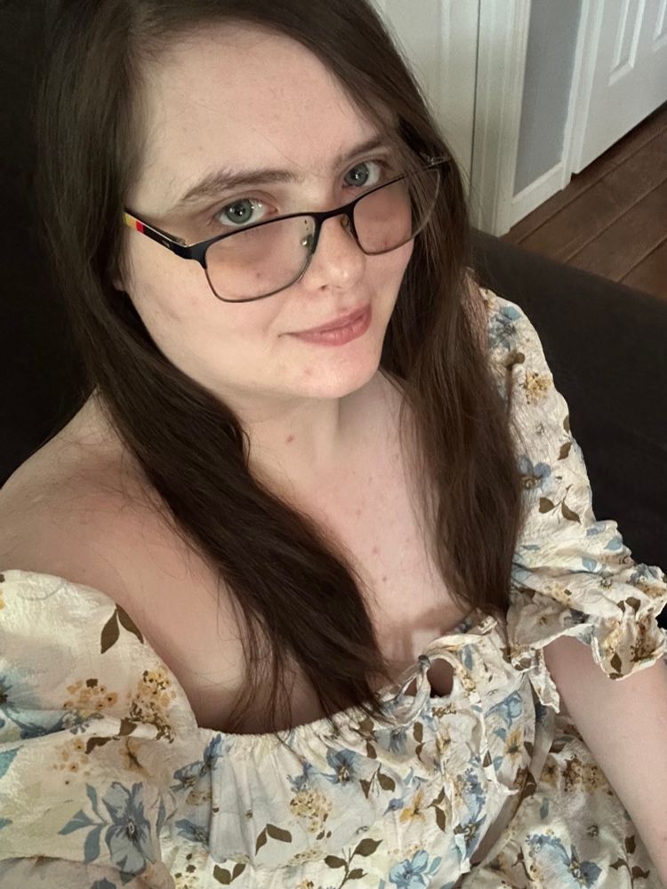 A girl with brunette hair and blue eyes wearing a pair of black glasses with rainbow arms and a floral dress that has blue and yellow flowers.