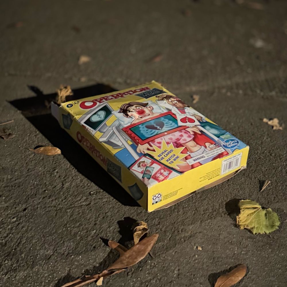 Picture of the game "operation" in a box on the sidewalk at night, with leaves and a harsh shadow on the top left