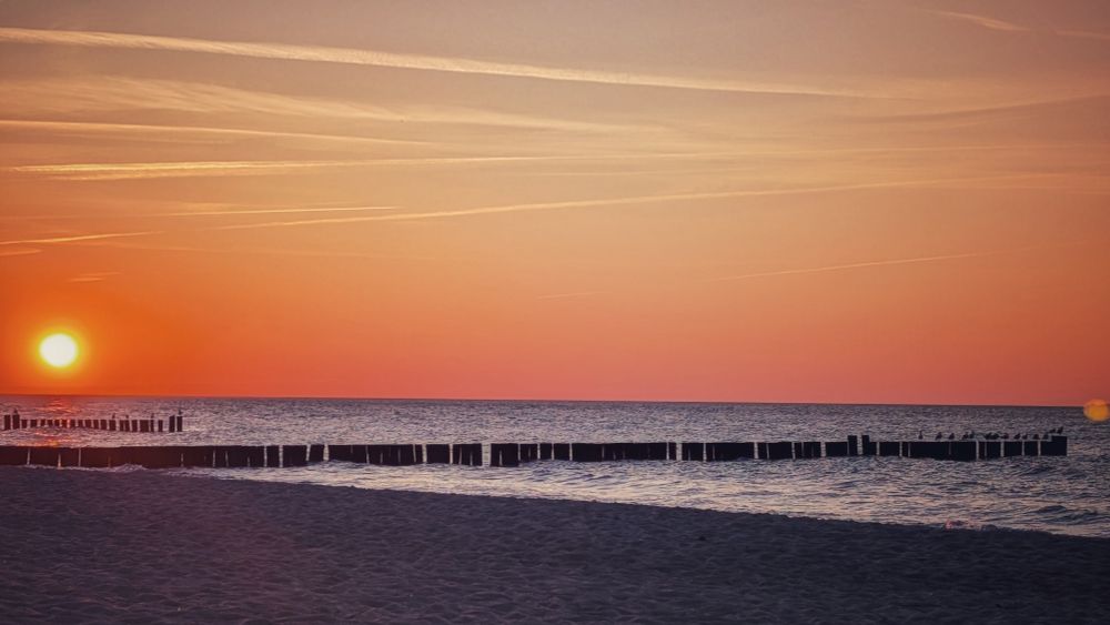 Sonnenuntergang am Meer. Links die Sonne als roter Ball kurz über dem Wasser. Im Vordergrund Strand, Holzsteelen reichen als Buhnen weit ins Wasser. Wolkenloser Himmel.