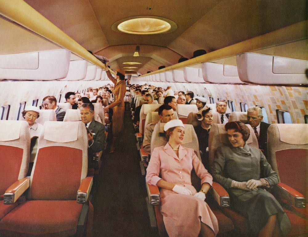 A photo from the 1970s of the inside of a commercial Boeing 707. Everyone is well-dressed in suits and dresses. Several of the women are wearing hats. A flight attendant (perhaps a stewardess, since it's the '70s) is in the aisle reaching towards an overhead shelf which appears to contain several men's hats.

There is not a single person of color in the photo, even though there are at least a hundred people in the photo.