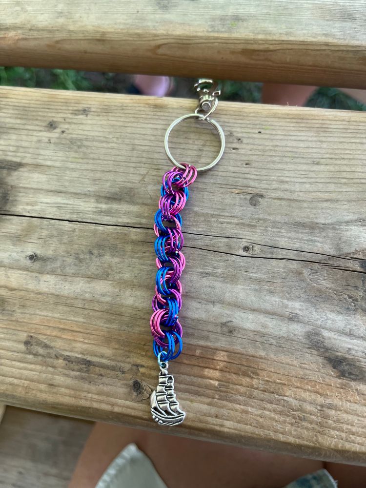 a chainmail keychain made in the colour of the bisexual flag with a ship charm hanging from the bottom.