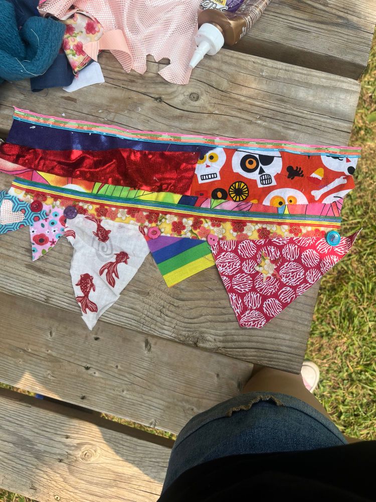 a picture of a little flag made out of various scraps of fabric. there are 5 triangles hanging from the bottom.Some of the scraps are:
fish pattern, rainbow, pirate pattern, various floral fabrics, 5 buttons in the middle spread throughout the flag, with two different colours of striped ribbon running through the middle and on the very top. there is a cut out heart on the left side, in one of the triangles, and a cut out star on the far-right side on one of the triangles.
