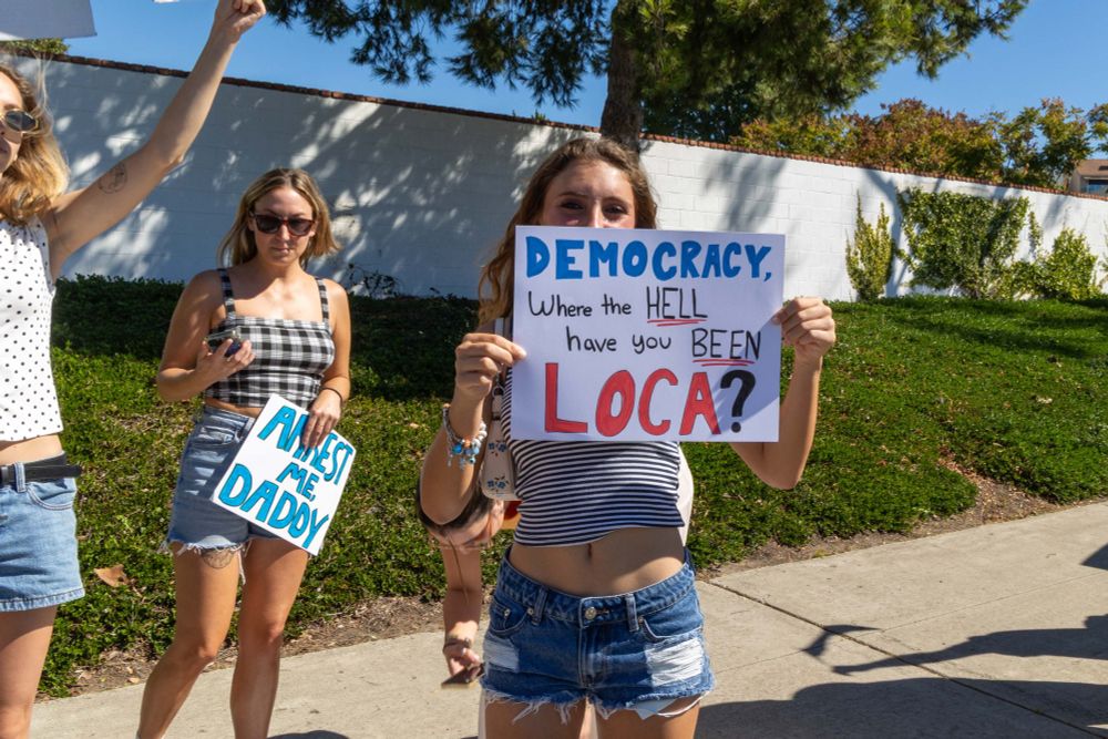 A protester at a No Kings protest holding a sign that says, "Democracy, Where the HELL have you BEEN? LOCA?"