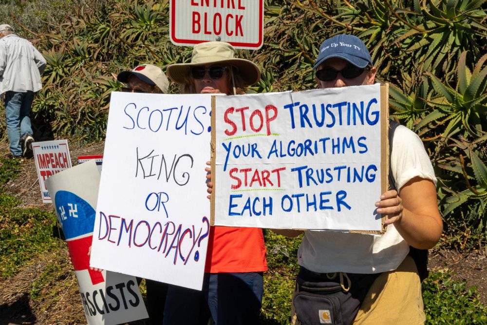 Protesters holding signs at a No Kings Protest. One sign says, "Scotus King or Democracy?" The other sign says, "Stop Trusting Your Algorithms And Start Trusting Each Other." 