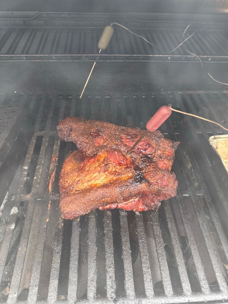 A photo of a pork shoulder on the bbq. A meat probe is sticking out and it has a dark bark
