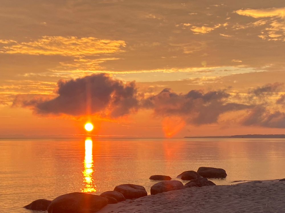 Sunset golden ball reflects on the lake beneath clouds. Rocks hug the shoreline in the water.