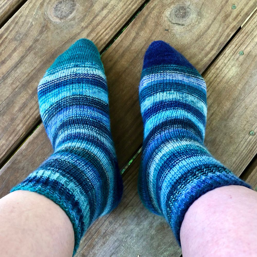 My bonus pair of socks on my feet, which are side by side on the deck floor.  My toes are pointing towards the top of the photo, teal on the left, blue on the right.  I really enjoy the way these socks go together without matching.  The stripes in both sock kits alternate light and dark, and I tried to keep them out of phase so that I would have light on light, light on dark, and darks on dark.  The different numbers of colors in the two kits (eight blue-green, six blue) also naturally shuffled things up.  The result is that in place of the crisp, four-to-five row stripes each ball of yarn makes on its own, these socks have broader stripes of mixed shades with blurrier edges.
