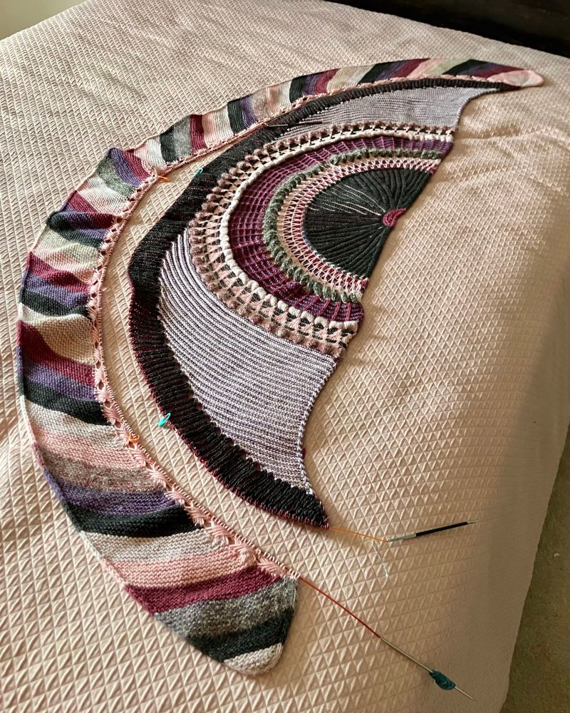 My Shawlography lying on a bed, midway through the process of grafting it back together. The color way consists of black, dark purple, dark cherry, flamingo pink, and purplish ecru. In the center of the semicircle, the shawl starts with a tiny cherry semicircle, surrounded by a fan of textured stripes in prominent black with the other colors peeking out from behind. The shawl design has a two-color brioche strip many sections later that echoes that central fan, with black in the raised foreground and cherry behind it. I flipped them, thinking the brighter color would look better on top, and my fervent desire to go back to the original design is what moved me to pick the shawl apart in the first place.  I thought I was going to knit a new startup row, attach the brioche strip the other way, and then make a second incision to flip the border, but I decided it was easier to knit a new brioche section from scratch so I only grafted once.