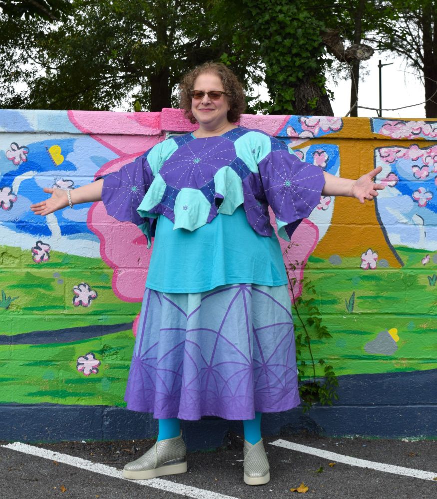 Me wearing the look I submitted to the 2025 Math + Fashion show at Bridges Eindhoven. From top to bottom: a purple, blue, and teal polygonal quilted top I machine-sewed and hand embroidered, an aqua camisole, a two-thirds-circle skirt printed with a hyperbolic tiling outlined in purple that fades from teal to purple, aqua tights, white and silver platform shoes by United Nude. I’m also wearing a bead crochet bracelet in white, green, blue, and purple.  My arms are spread wide to show off the bell sleeves of the top.  Behind me is part of a mural by Carrie Patterson, Ash Scherer, and community art students from St. Mary’s College of Maryland.

Spoonflower printed the fabric for the top and the skirt.  The polygons for the top are printed on cotton jersey fabric, while the skirt panels and the fabric for the waist and pockets are on a cotton/linen blend.